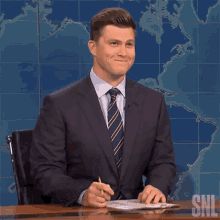 a man in a suit and tie is sitting at a desk with a snl logo on the wall behind him