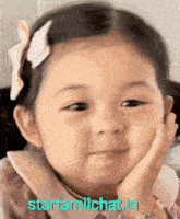 a little girl with a bow in her hair is smiling with her hand on her chin ..
