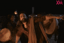a man with a beard is dancing in front of a group of drummers with xhk written in red