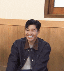 a man in a black jacket is smiling in front of a wooden wall .
